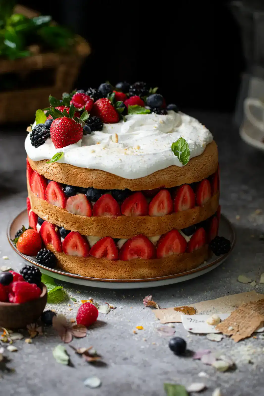 A layered sponge cake filled with fresh strawberries, blueberries, blackberries, and cream, topped with whipped cream and assorted berries, on a gray surface.