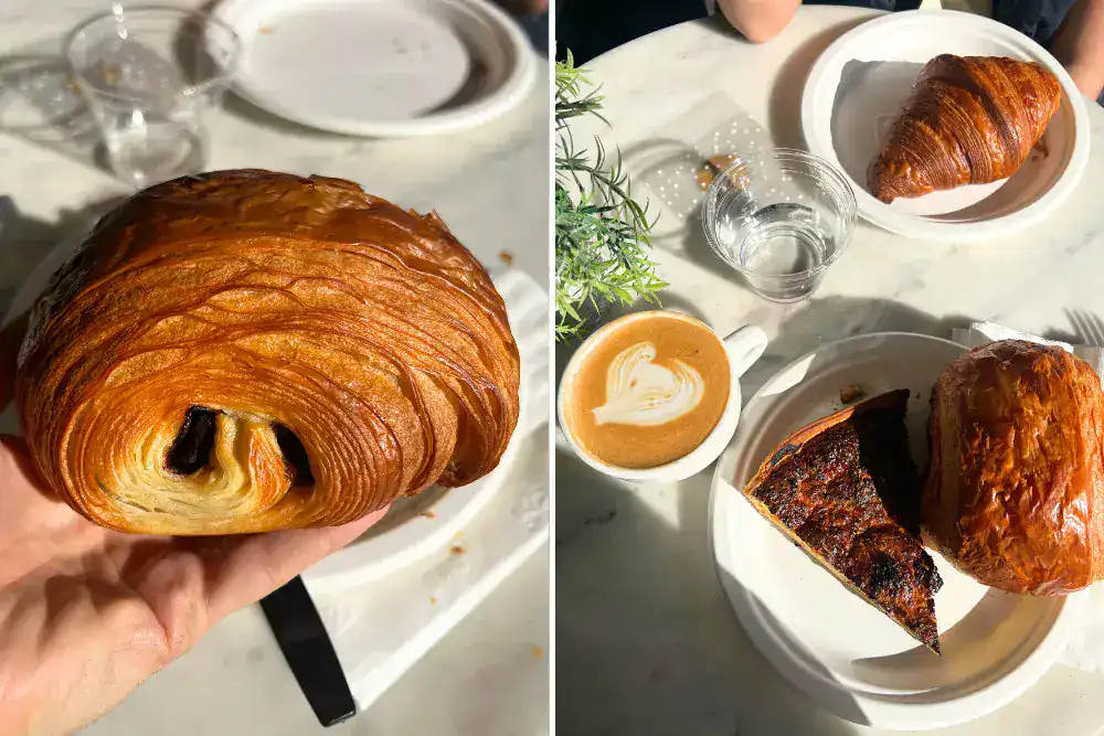 A hand holds a pain au chocolat. Next to it, a table with a croissant, water glass, a cappuccino, and a slice of quiche on a white plate.