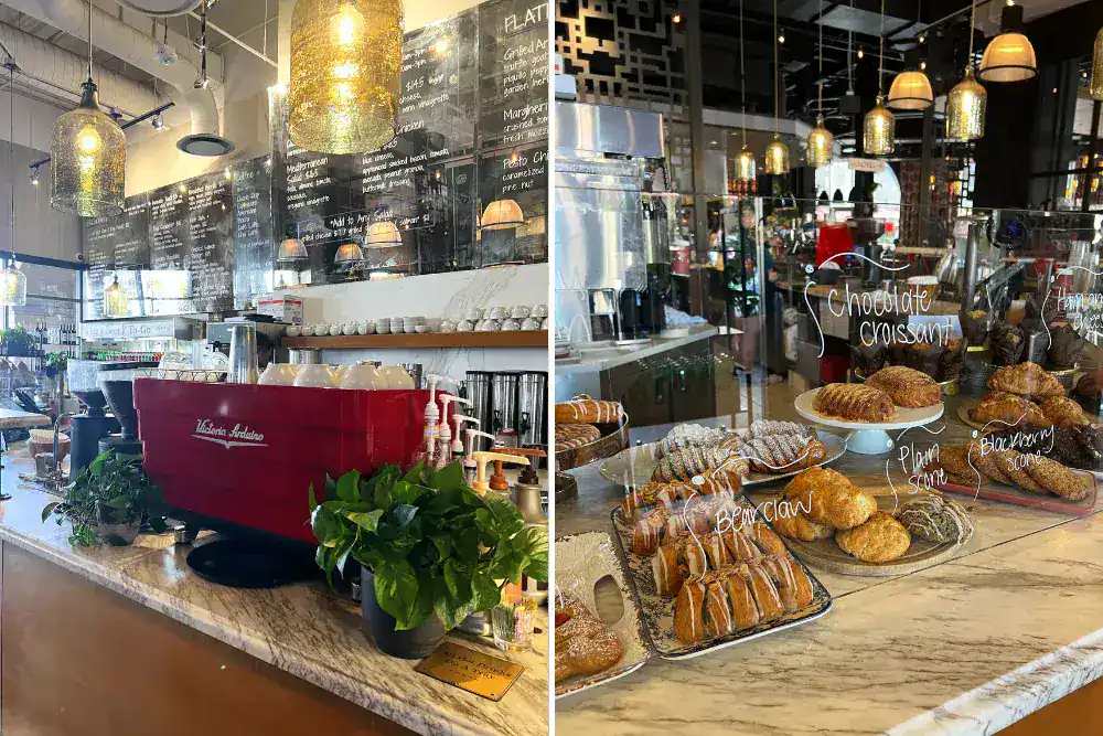 A modern café interior with a marble counter, coffee machine, hanging lights, menu boards, and a display of assorted pastries labeled with handwritten signs.