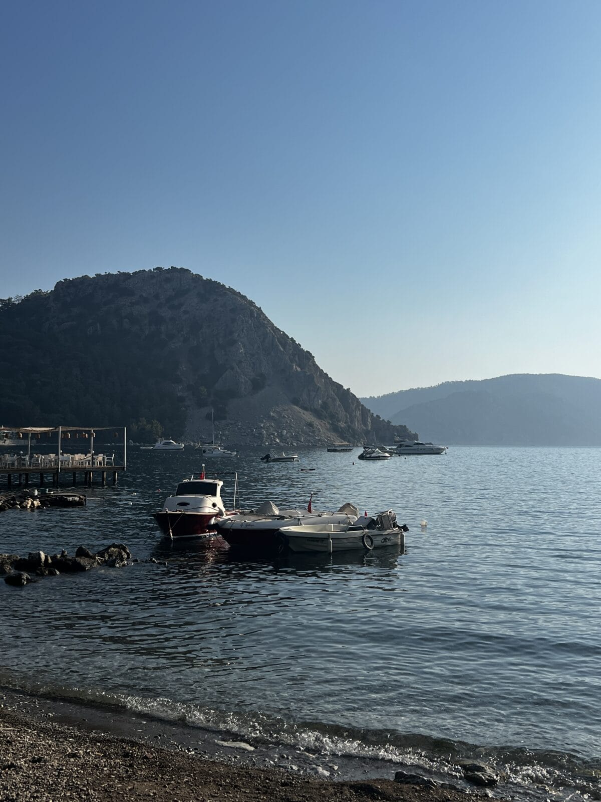 Small boats are docked near a rocky shore with calm water, a wooden pier, and tree-covered hills in the background under a clear blue sky.