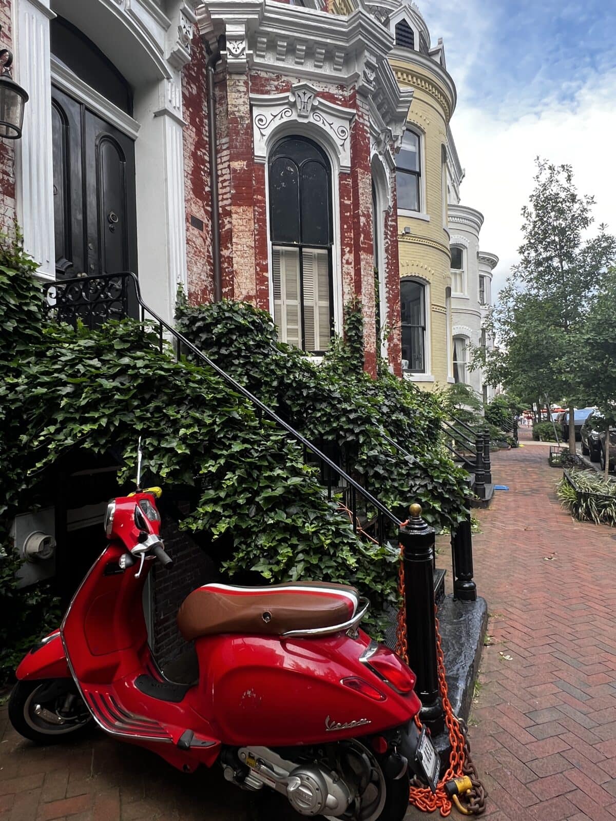 A red Vespa scooter is parked and chained outside a brick building with ivy-covered stairs on a city sidewalk.