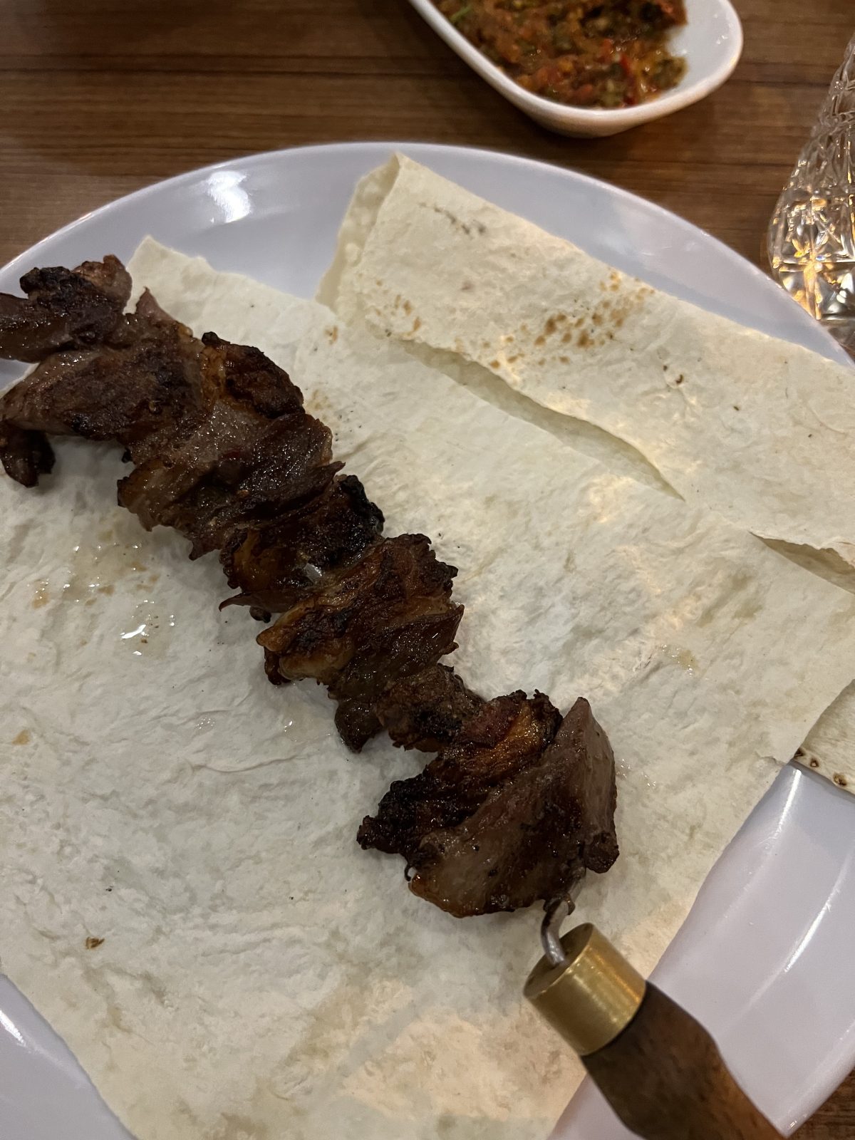 A skewer of grilled meat placed on flatbread, served on a white plate with a glass of water and a small dish of sauce in the background.