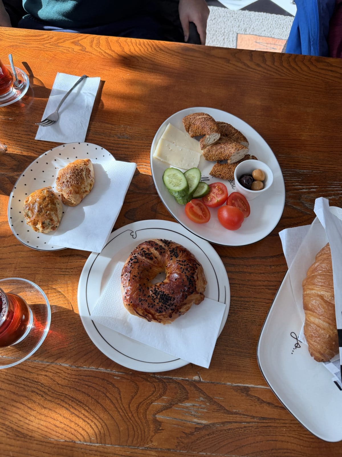 A wooden table set with tea, a bagel, a croissant, two scones, and a plate of cheese, falafel, cucumber, tomato, and olives.