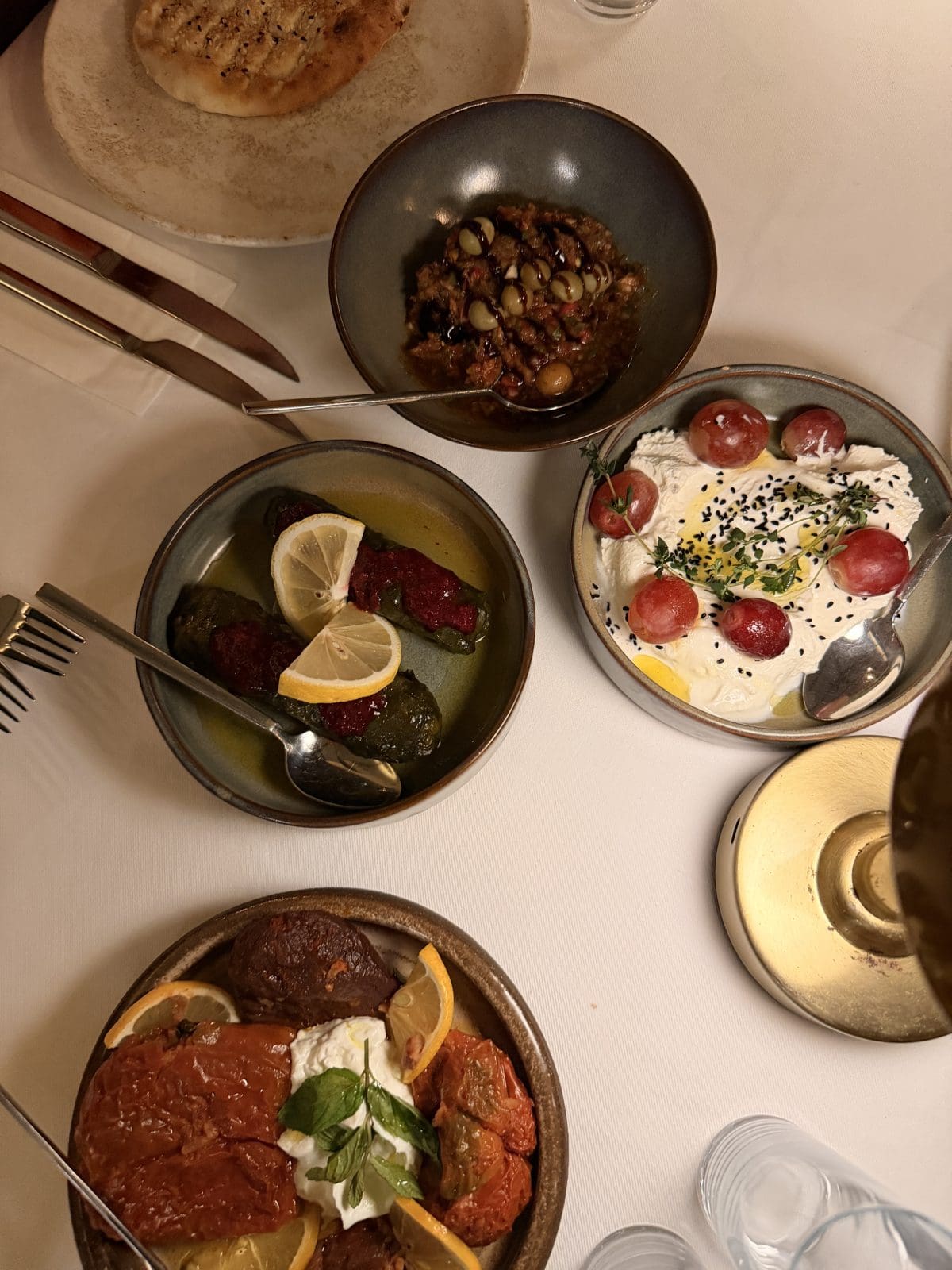A table set with Mediterranean dishes including stuffed grape leaves with lemon, whipped cheese with tomatoes, stewed vegetables, and a bowl of olives and tapenade, next to bread.