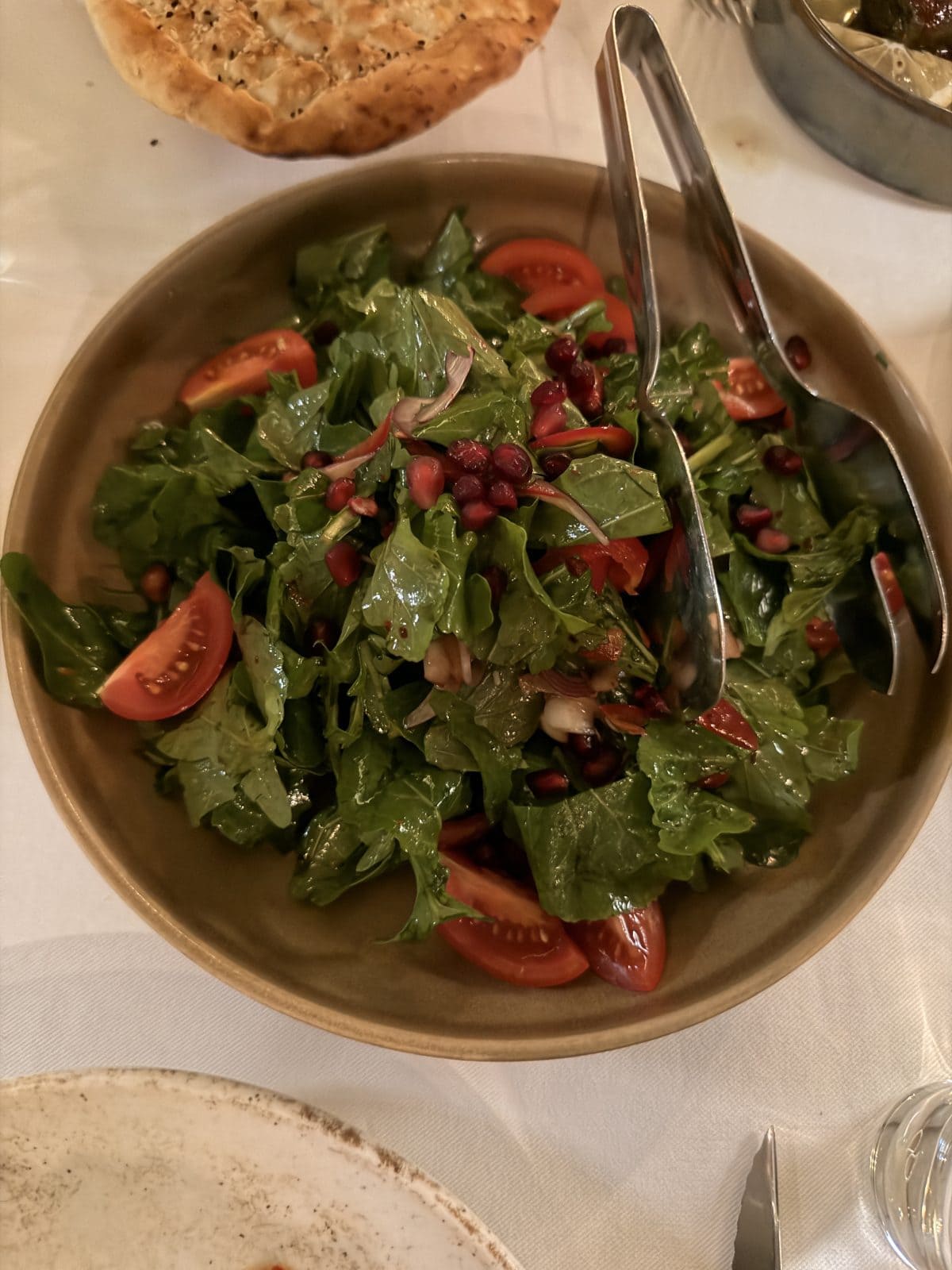 A bowl of fresh salad with arugula, tomato wedges, pomegranate seeds, and sliced onions, served with metal salad tongs on a table.