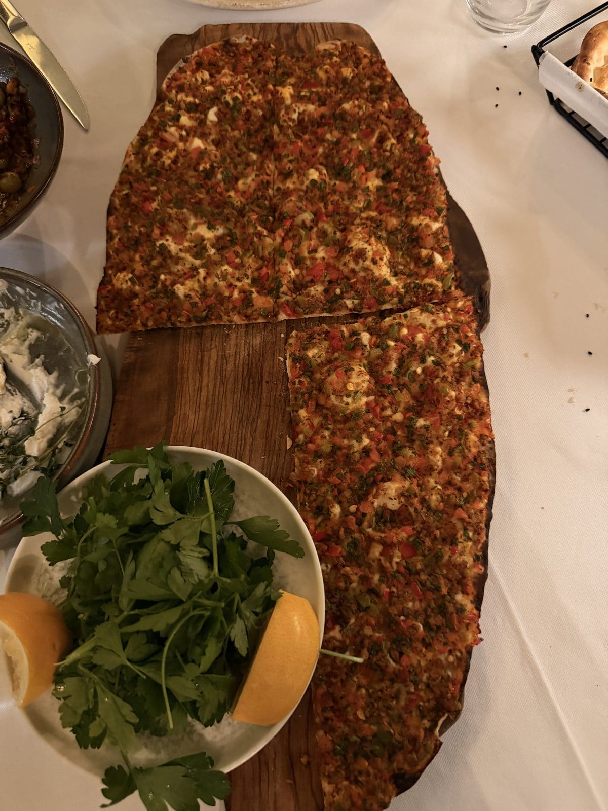 A rectangular flatbread topped with minced meat and vegetables on a wooden board, served with a plate of fresh parsley and lemon wedges.