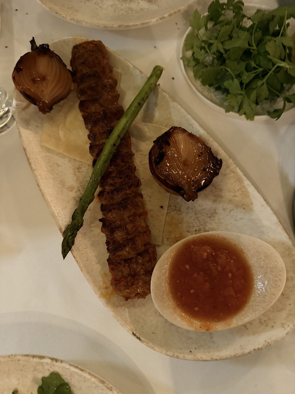A plate with a grilled kebab, a single asparagus spear, two grilled onion halves, a side of red sauce in a small bowl, and a plate of fresh herbs.