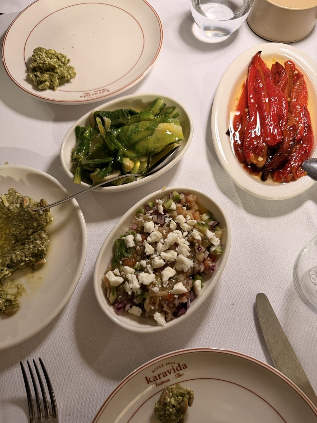 A table set with plates of assorted appetizers, including roasted red peppers, a cheese and vegetable salad, green beans, and a green spread, on a white tablecloth.