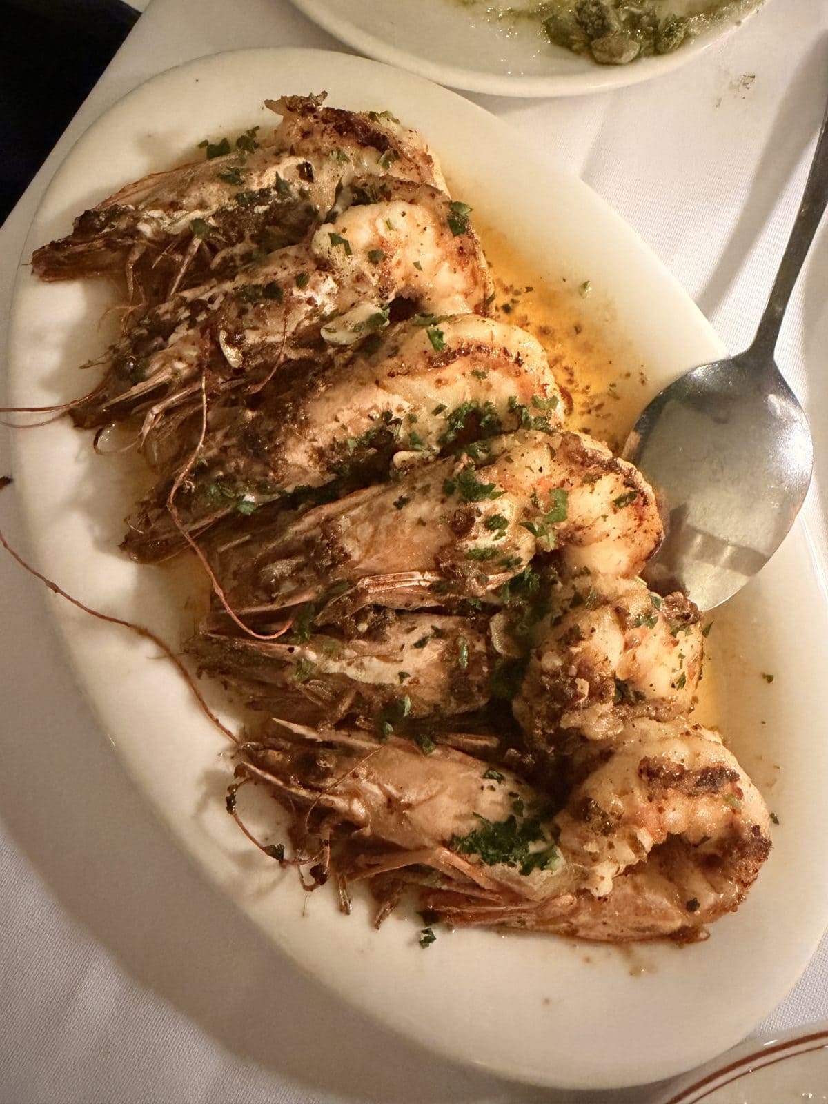A white platter with grilled shrimp in garlic butter sauce, garnished with chopped parsley, and a serving spoon on the side.