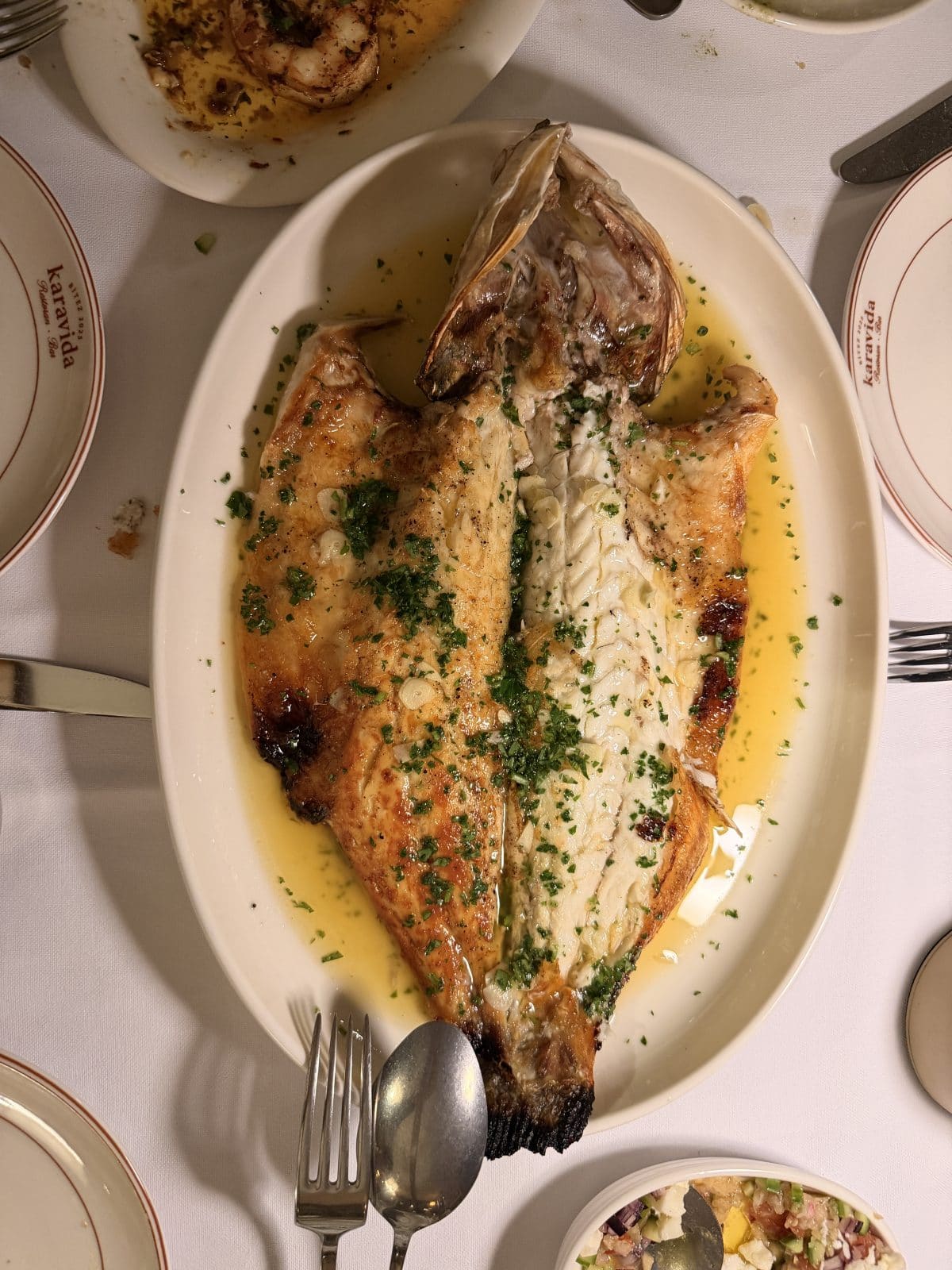 A whole grilled fish, split and garnished with chopped parsley and sauce, is served on an oval plate with utensils on a white tablecloth.