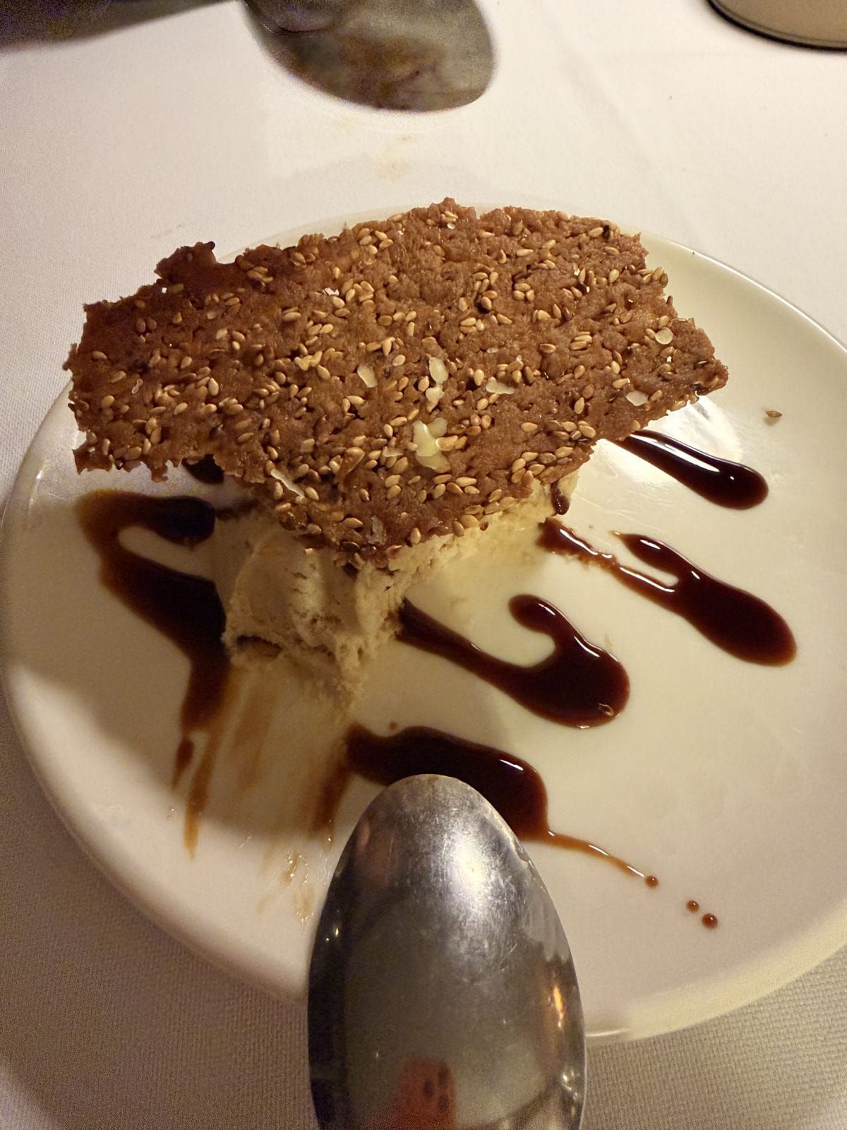 A dessert featuring a scoop of ice cream topped with a large sesame seed crisp, served on a white plate with chocolate sauce drizzle, and a spoon in the foreground.