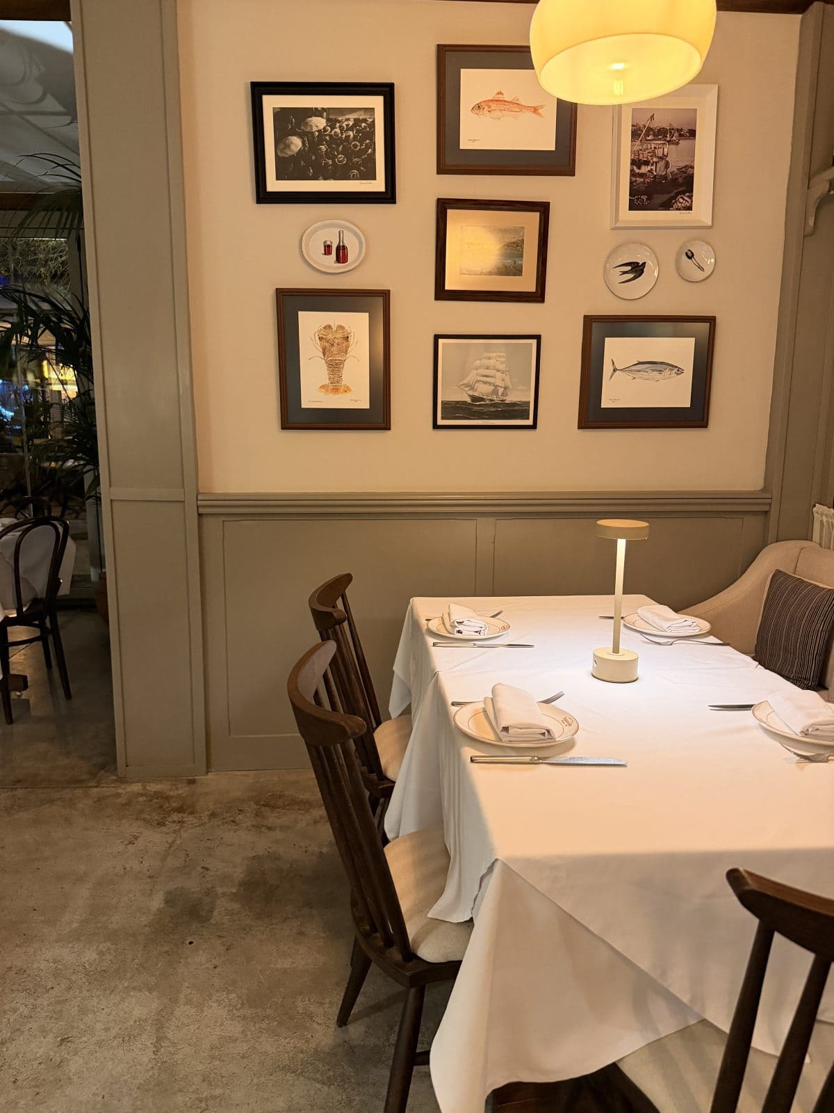 A neatly set dining table with wooden chairs, a white tablecloth, and a small lamp, positioned beside a wall decorated with framed pictures and plates.