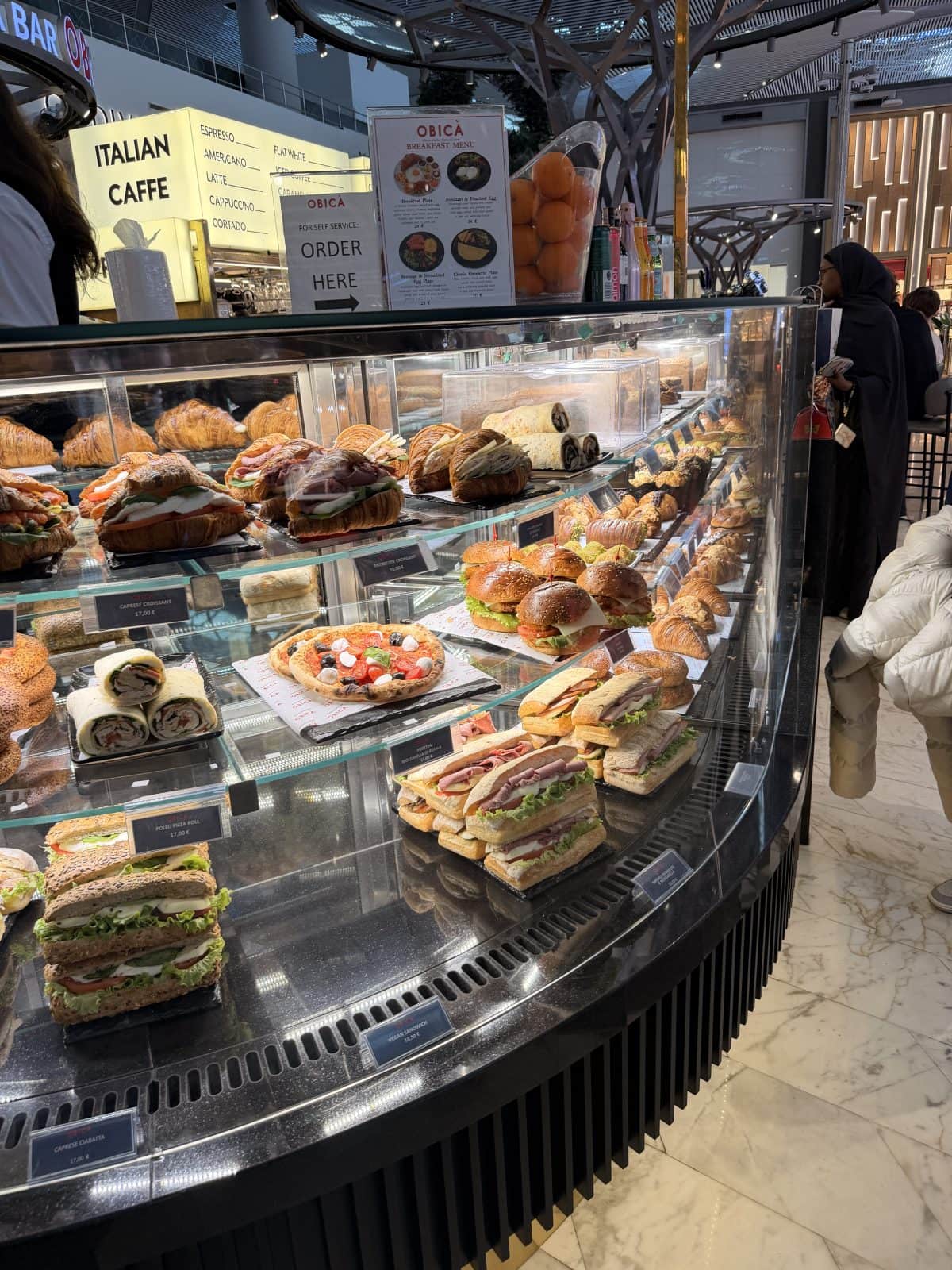 A glass display case filled with assorted sandwiches, pastries, and desserts in a modern cafe with people ordering in the background.