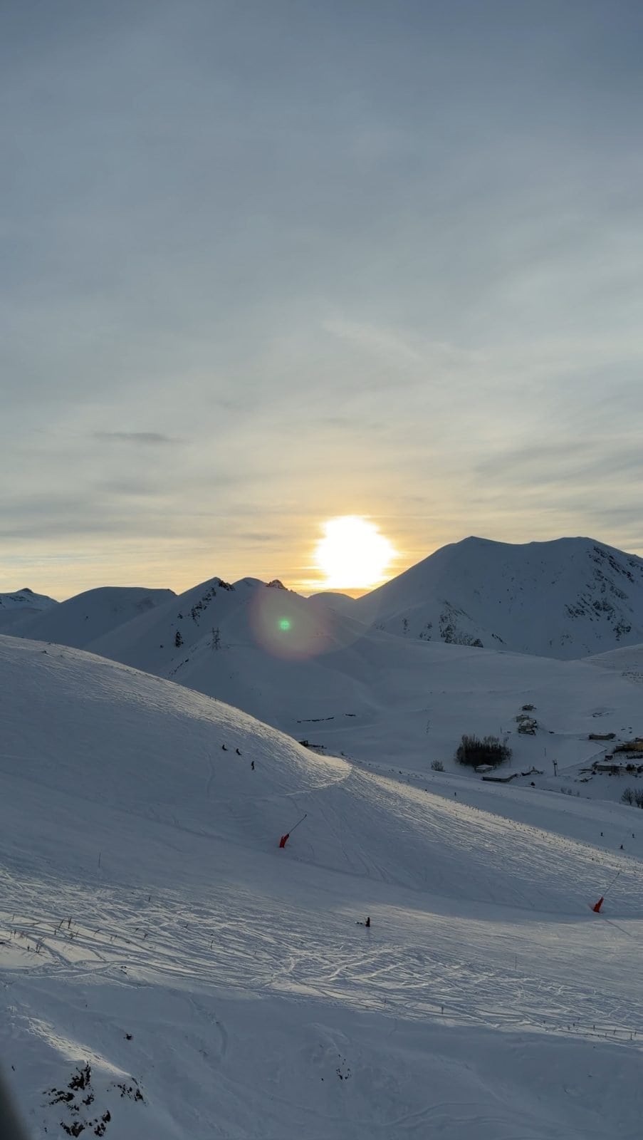 The sun sets behind snow-covered mountains near Erzurum, Türkiye, casting a soft light over a snowy landscape dotted with a few scattered skiers and ski tracks.