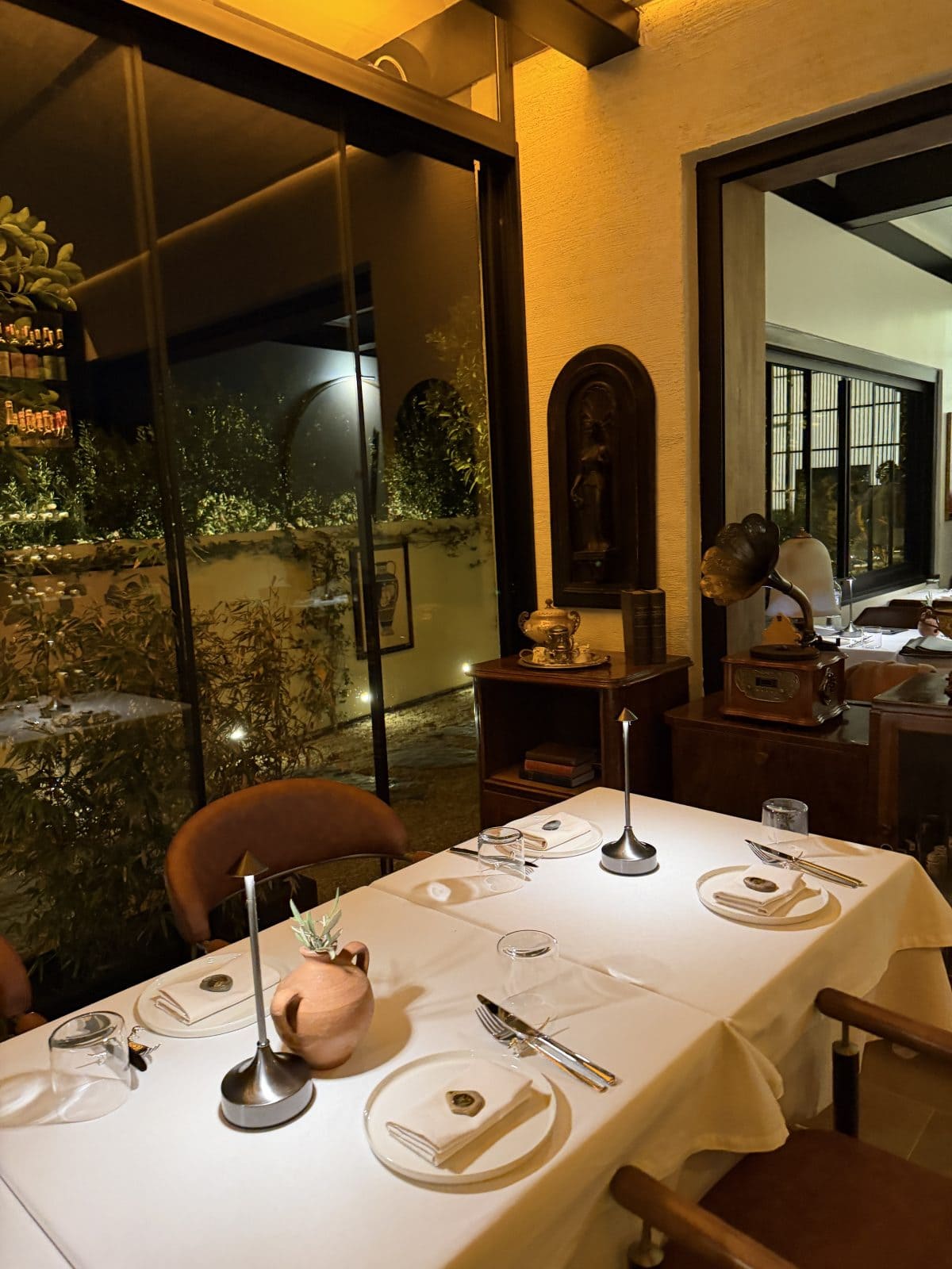 A dimly lit restaurant table set for four near a large window, with vintage decor and greenery visible outside.
