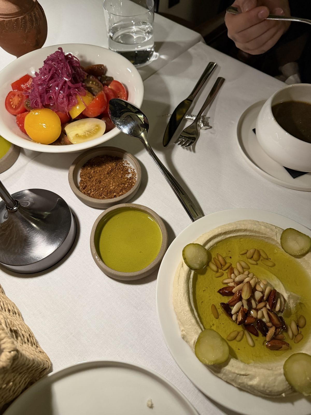 A table set with a bowl of colorful salad, a plate of hummus topped with pine nuts and pickles, olive oil, seasoning, a glass of water, soup, and cutlery.