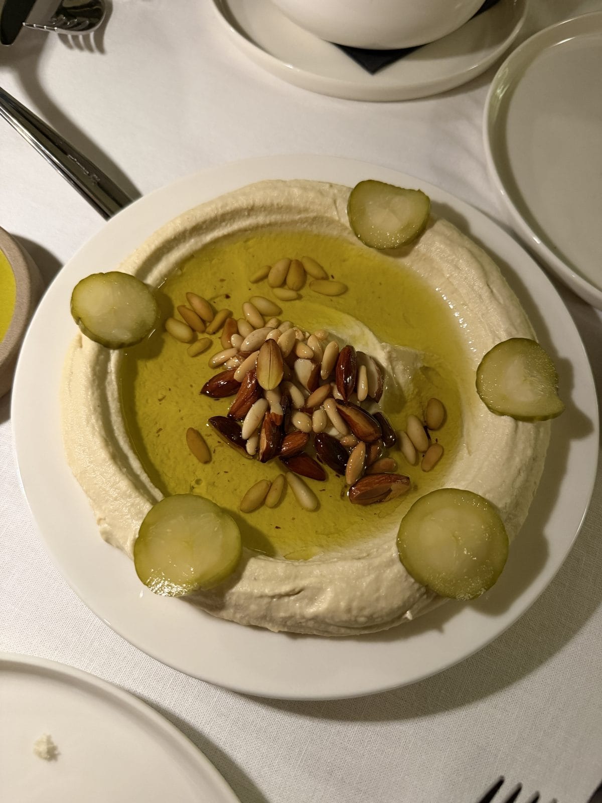 A plate of hummus topped with olive oil, pine nuts, almonds, and garnished with sliced pickles, served on a white tablecloth.
