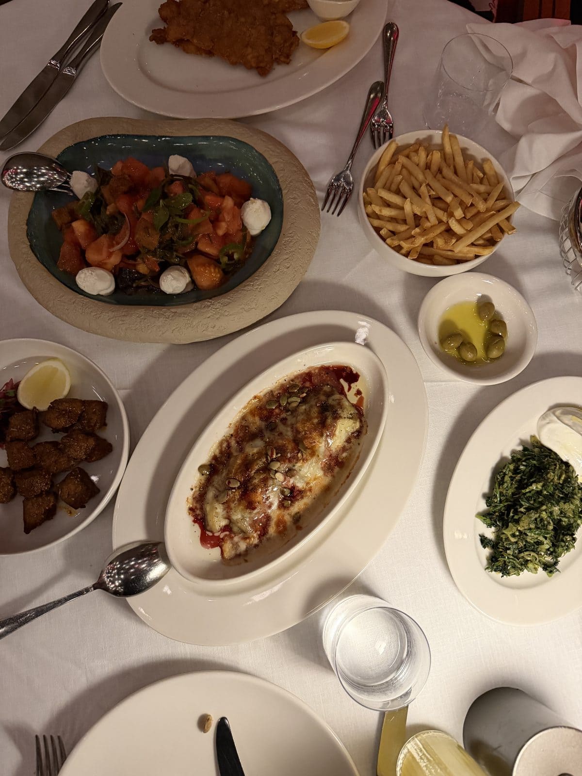 A table set with various dishes including baked cheese, sautéed greens, French fries, olives, a tomato salad with mozzarella, bread cubes, a cutlet, and drinks.