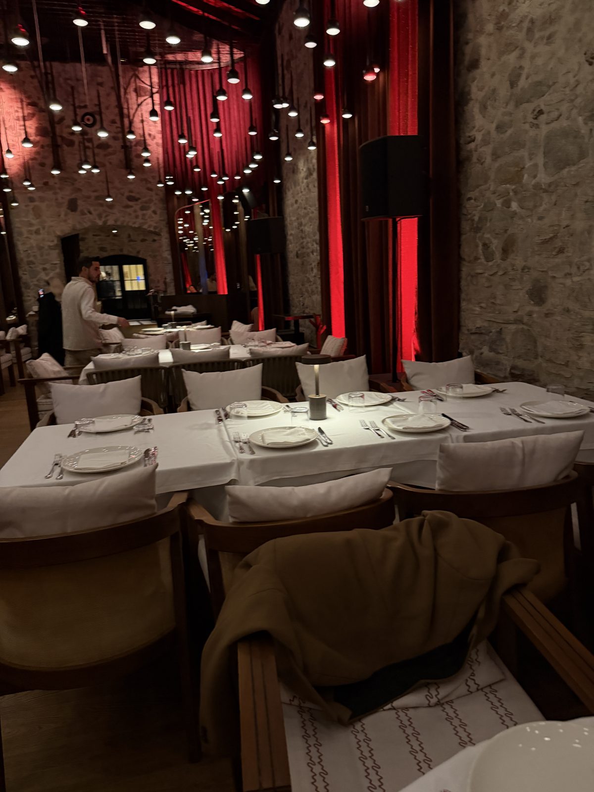 Dimly lit restaurant with stone walls, red accent lighting, and tables set with white plates and cloth napkins. A person stands near one table; a coat is draped over a chair in the foreground.