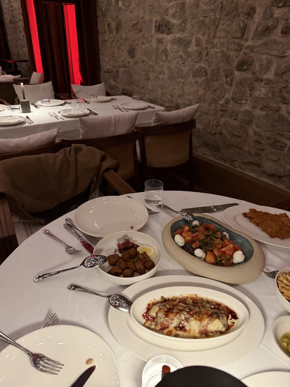 A restaurant table with plates of assorted dishes, including a baked dish, salad, fried food, meatballs, and sauces; empty tables and stone wall in the background.
