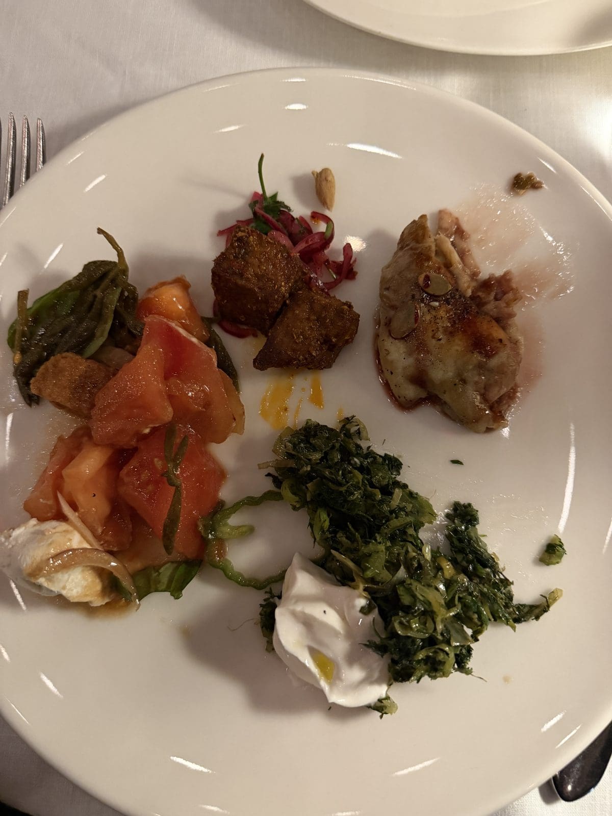 A white plate with small portions of cooked spinach, tomato salad, meat, fried cubes, red cabbage slaw, and a dollop of white sauce on a table.