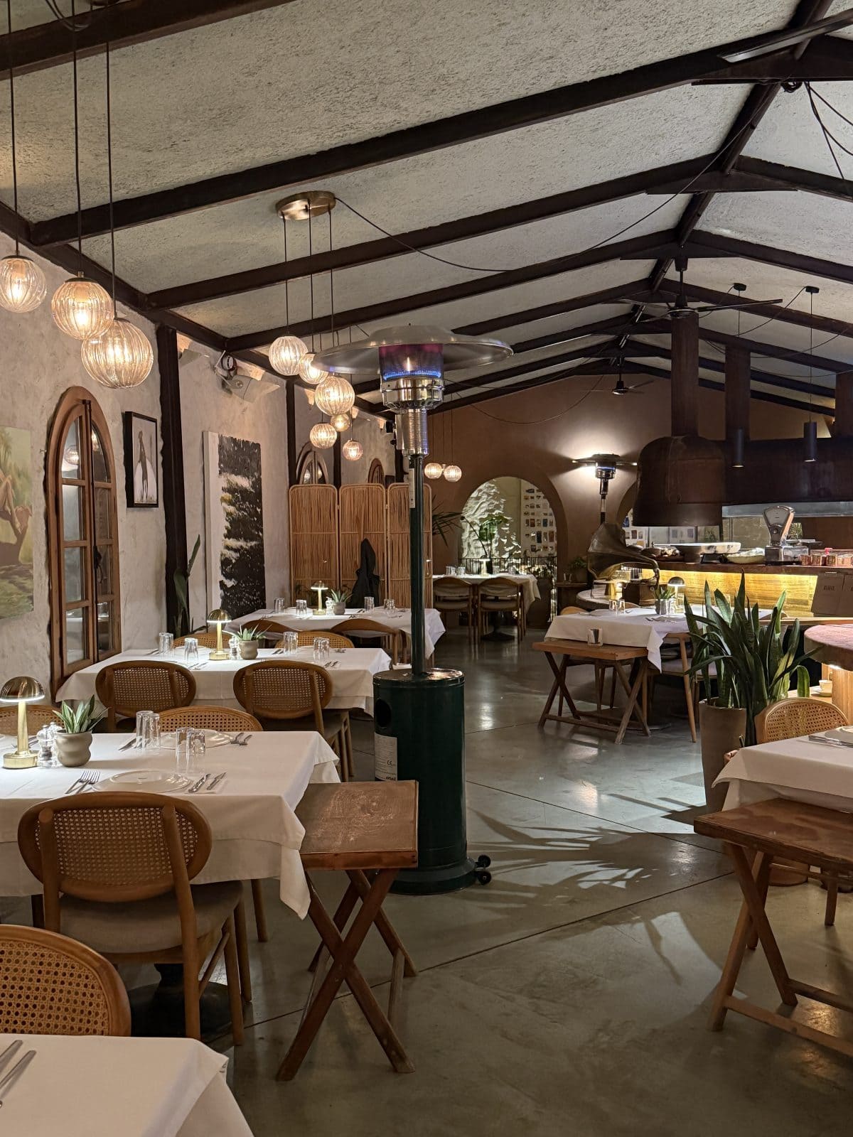 A cozy, warmly lit restaurant interior with neatly set tables, wooden chairs, hanging lights, potted plants, and an open kitchen in the background.