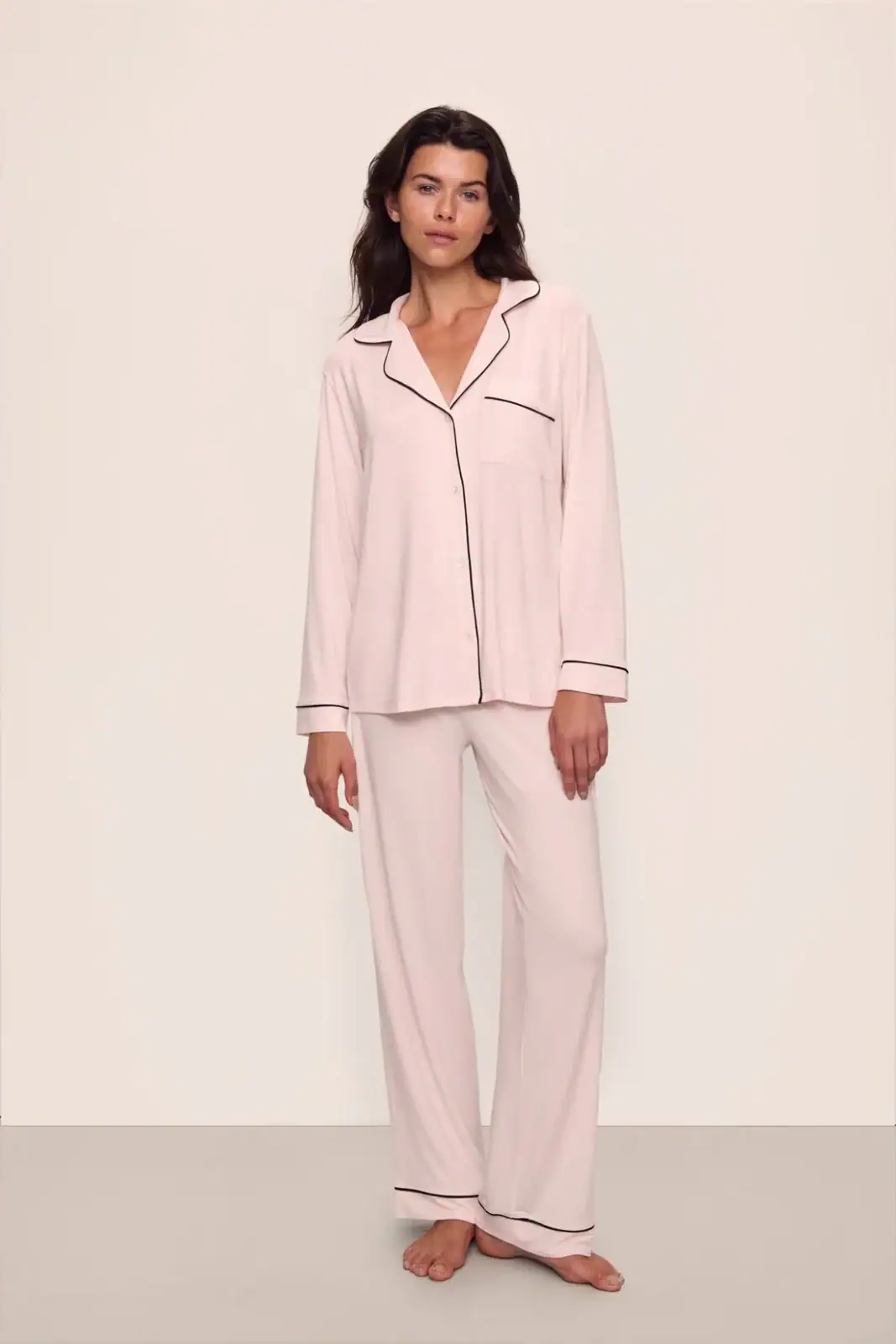 A person stands barefoot wearing a light pink pajama set with black piping, consisting of a long-sleeve top and loose pants, against a plain background.
