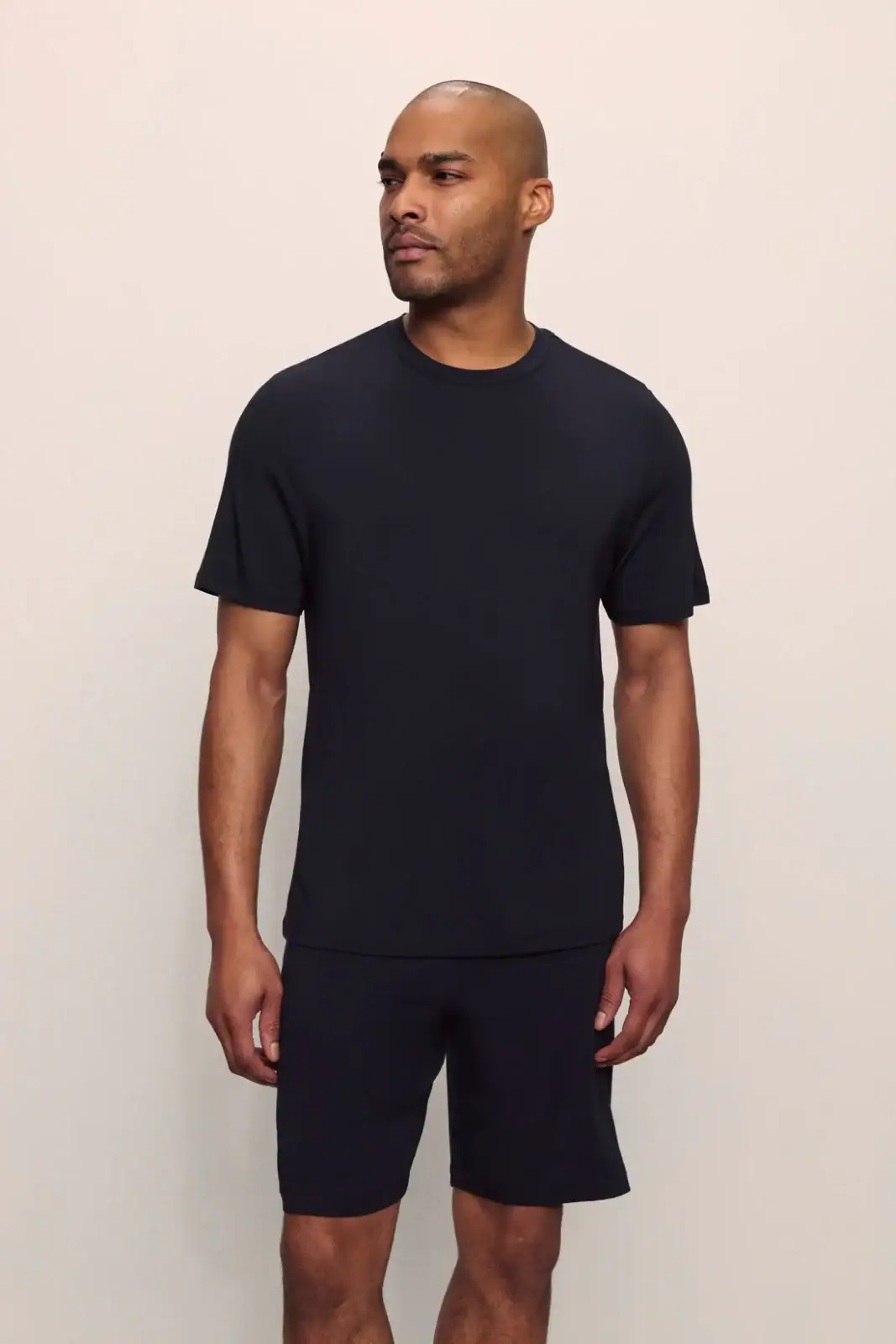 A man stands against a plain background, wearing a black short-sleeve t-shirt and black shorts.