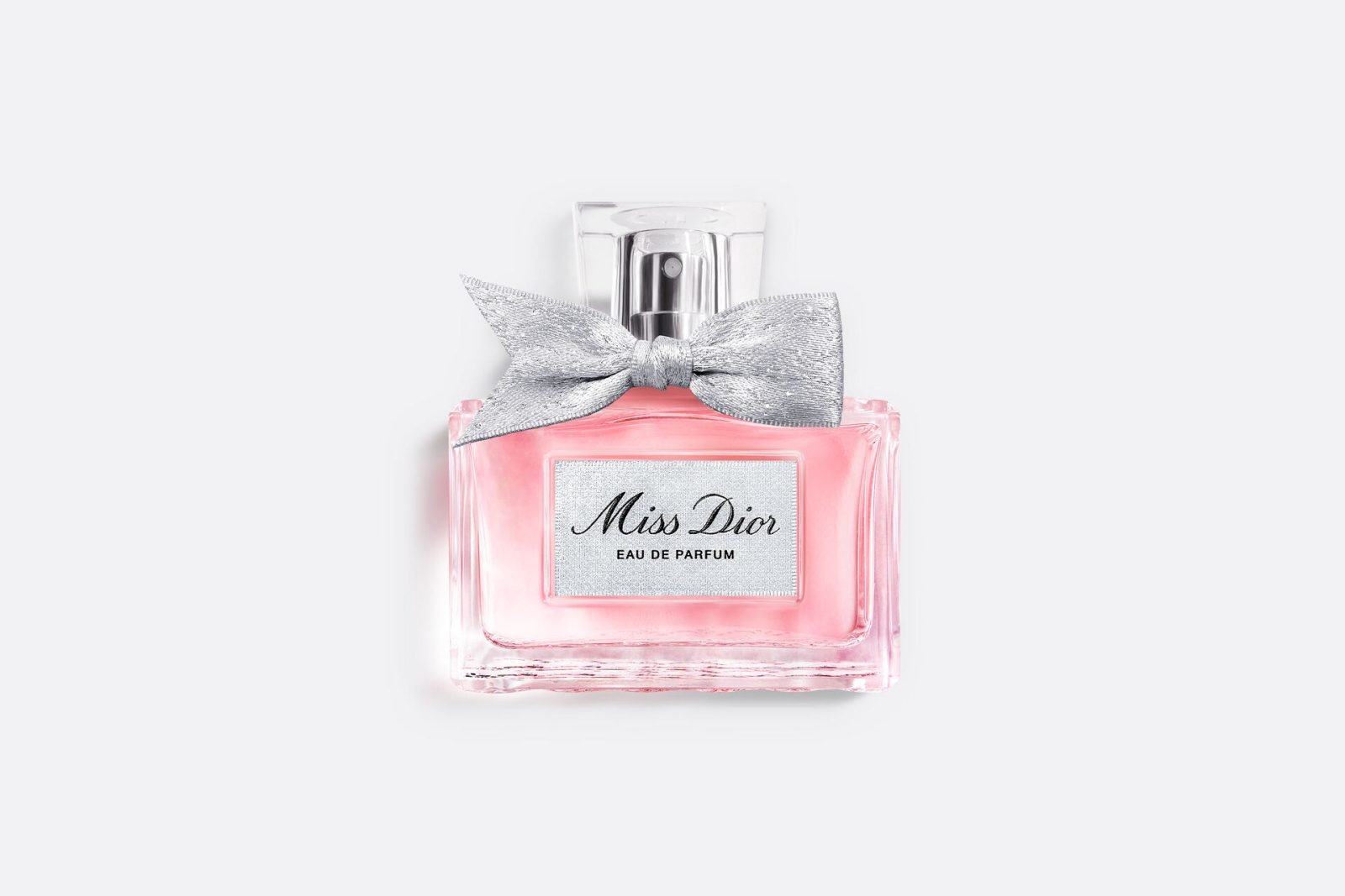 A clear glass bottle of Miss Dior Eau de Parfum with pink liquid inside, topped with a silver bow and a rectangular label on the front.