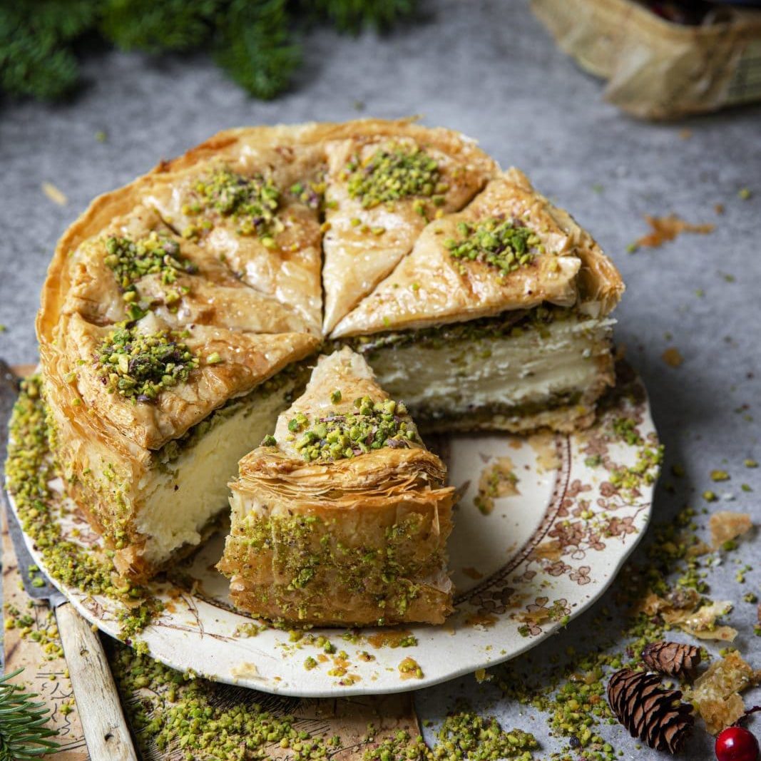A sliced piece of layered baklava cheesecake topped with chopped pistachios sits on a decorated plate, surrounded by festive greenery and berries.