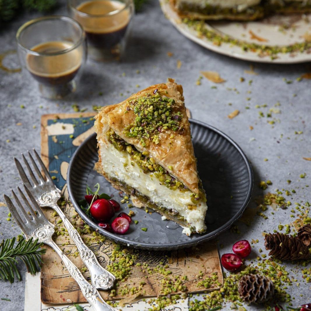 A slice of layered pastry dessert topped with chopped pistachios on a black plate, garnished with cranberries, beside two forks and glasses of coffee.
