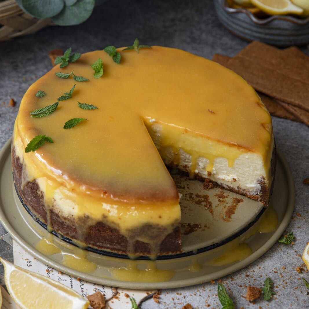 A cheesecake topped with yellow lemon glaze and mint leaves, with one slice removed. Fresh lemon slices and mint leaves are scattered around on the table.