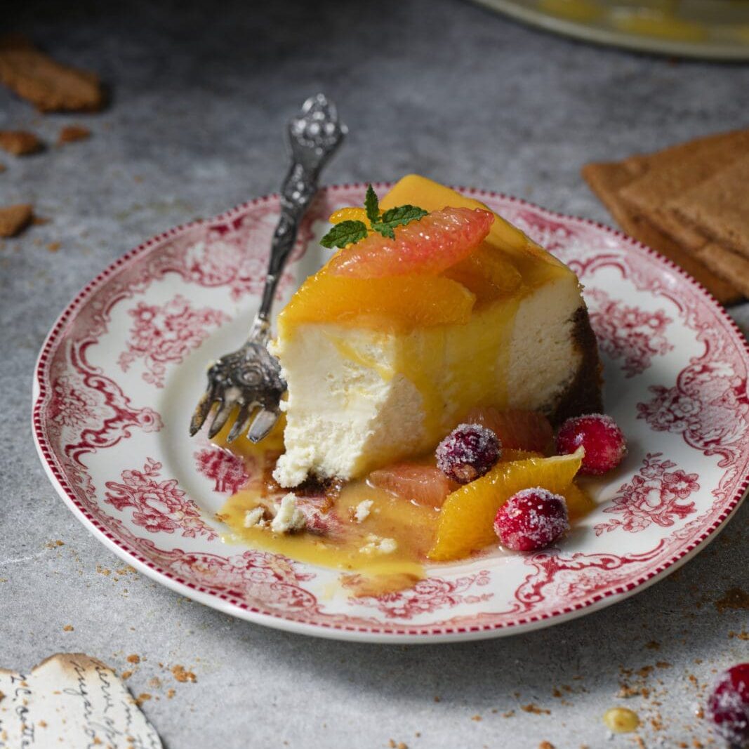 A slice of cheesecake with fruit topping and sauce is served on a decorative plate with a fork, surrounded by crumbs and garnished with mint and berries.
