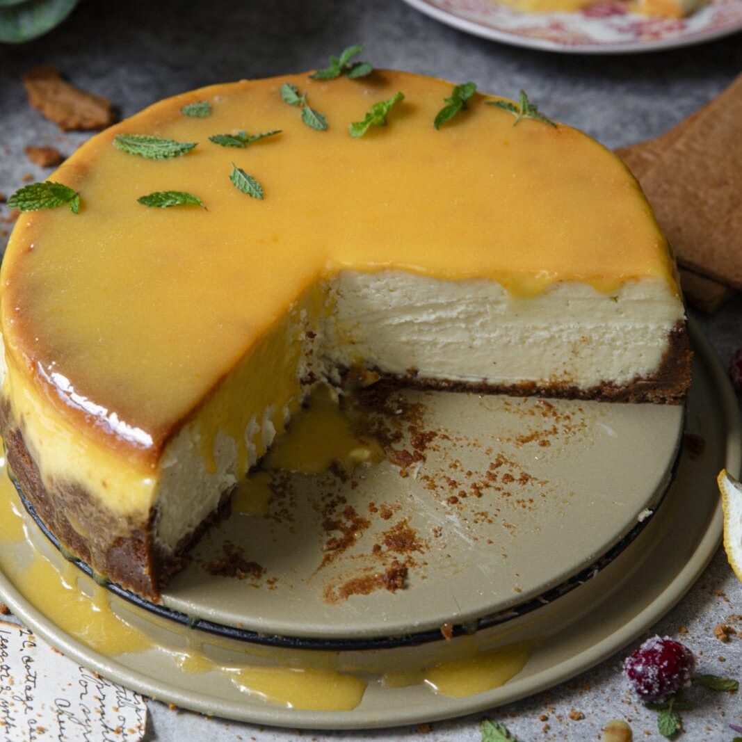 A cheesecake with a yellow glaze and mint leaves sits on a plate with a slice removed; crumbs and sauce are visible around the cake.