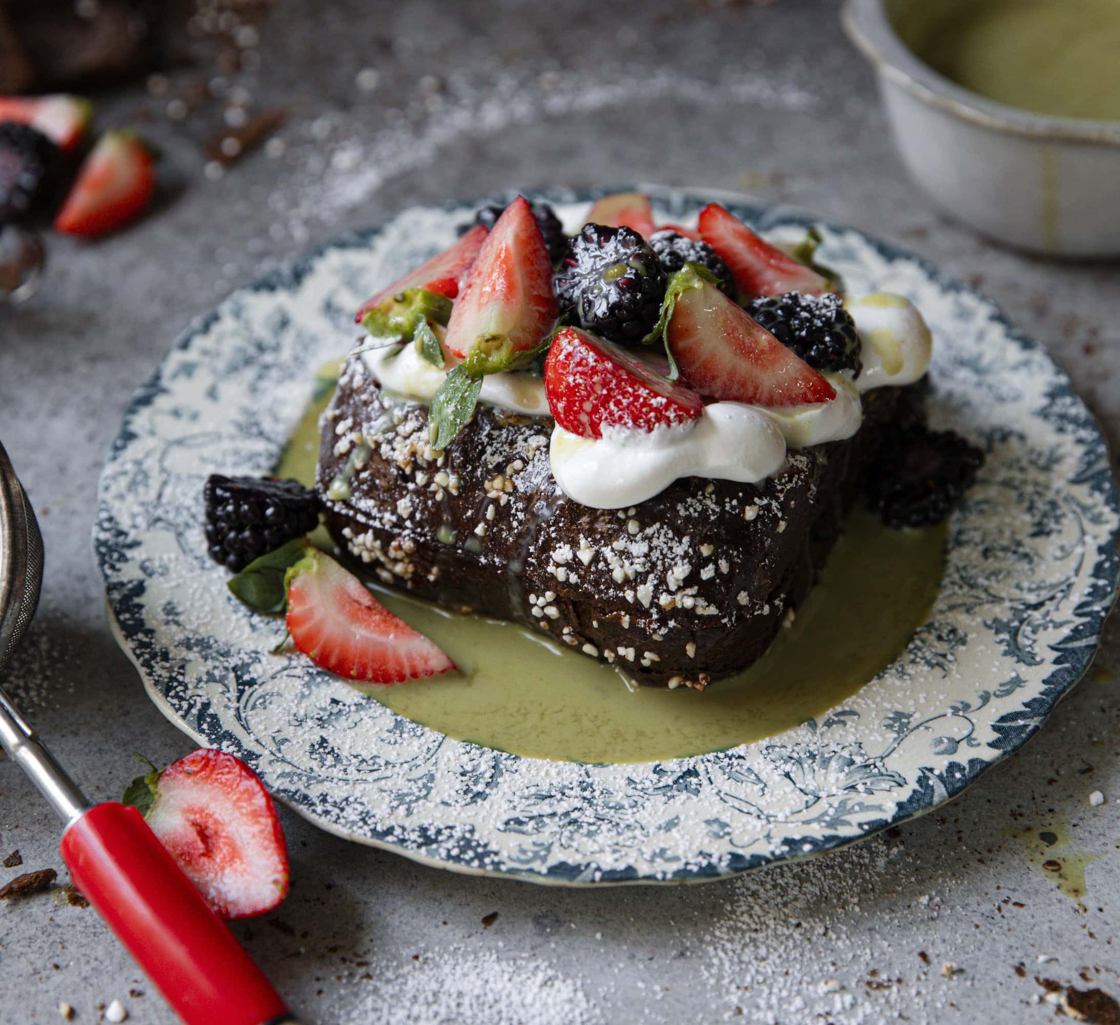 A slice of chocolate cake topped with whipped cream, sliced strawberries, and blackberries on a decorative plate, with a bowl of green sauce and cut strawberries nearby.