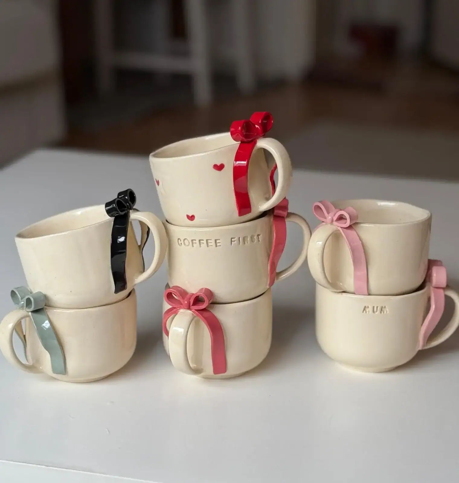 Six beige ceramic mugs with bows on their handles are stacked in two columns; two mugs have text, one says "COFFEE FIRST" and the other "MUM," while others are plain.