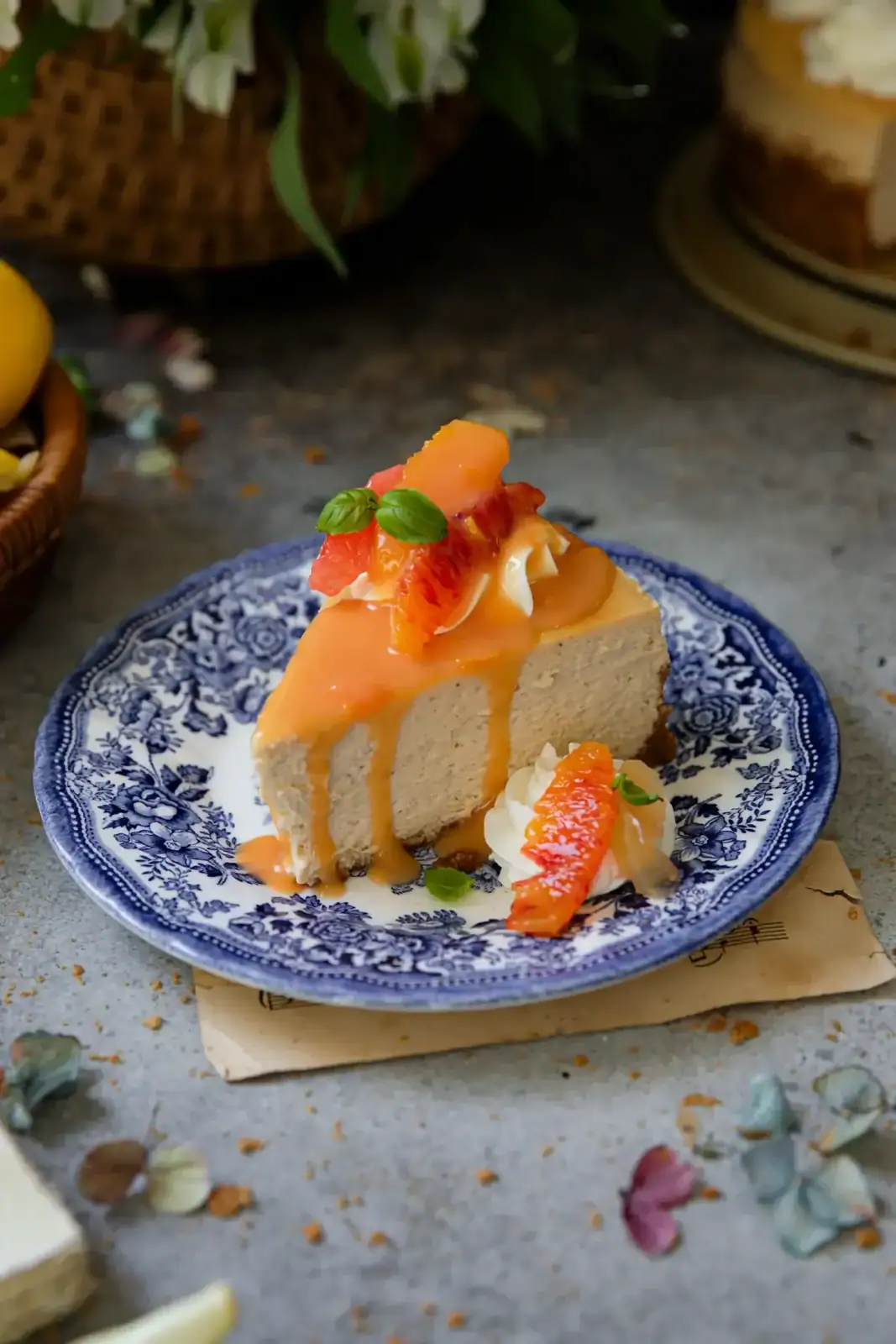 A slice of cheesecake topped with orange sauce, citrus segments, and fresh herbs on a blue and white patterned plate.