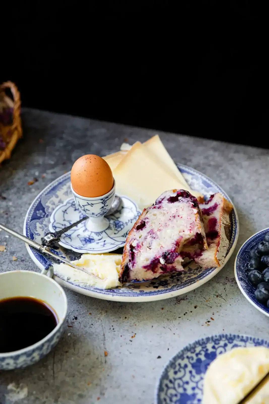 A plate with a soft-boiled egg in an egg cup, cheese slices, a slice of bread with blueberry spread, and a fork. A cup of coffee and a bowl of blueberries are nearby.