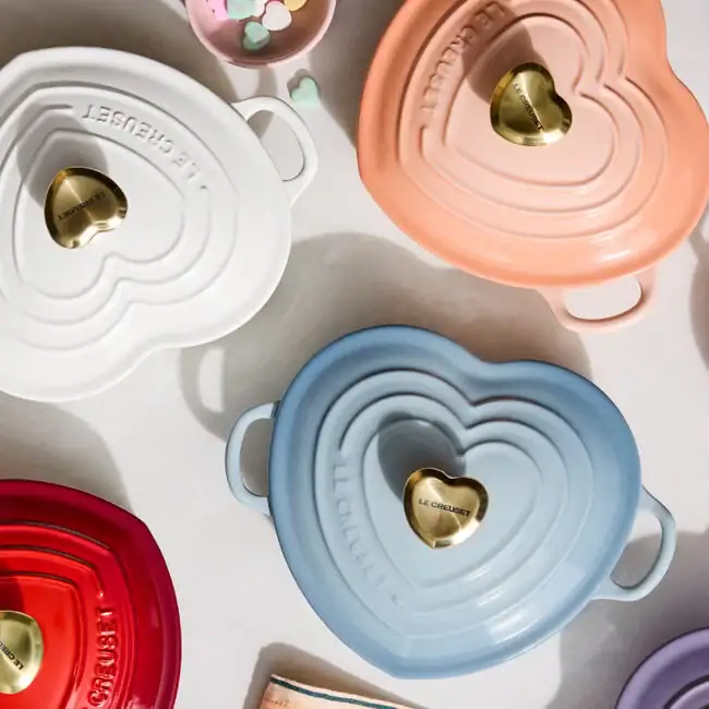 Assorted heart-shaped Le Creuset Dutch ovens in different colors, including white, peach, blue, and red, arranged on a light surface.